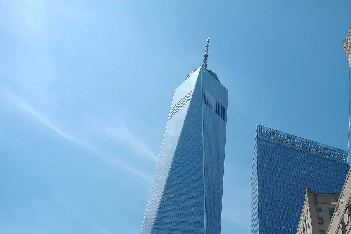 One World Trade Center, a tapering blue glass tower with a spire, seen from the street against a clear sky