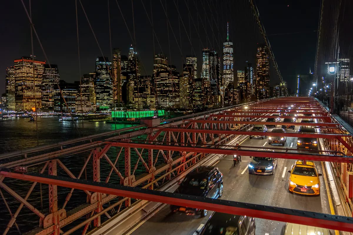 Night traffic and yellow cabs on the Brooklyn Bridge roadway beneath red steel trusses, with the lit Manhattan skyline beyond
