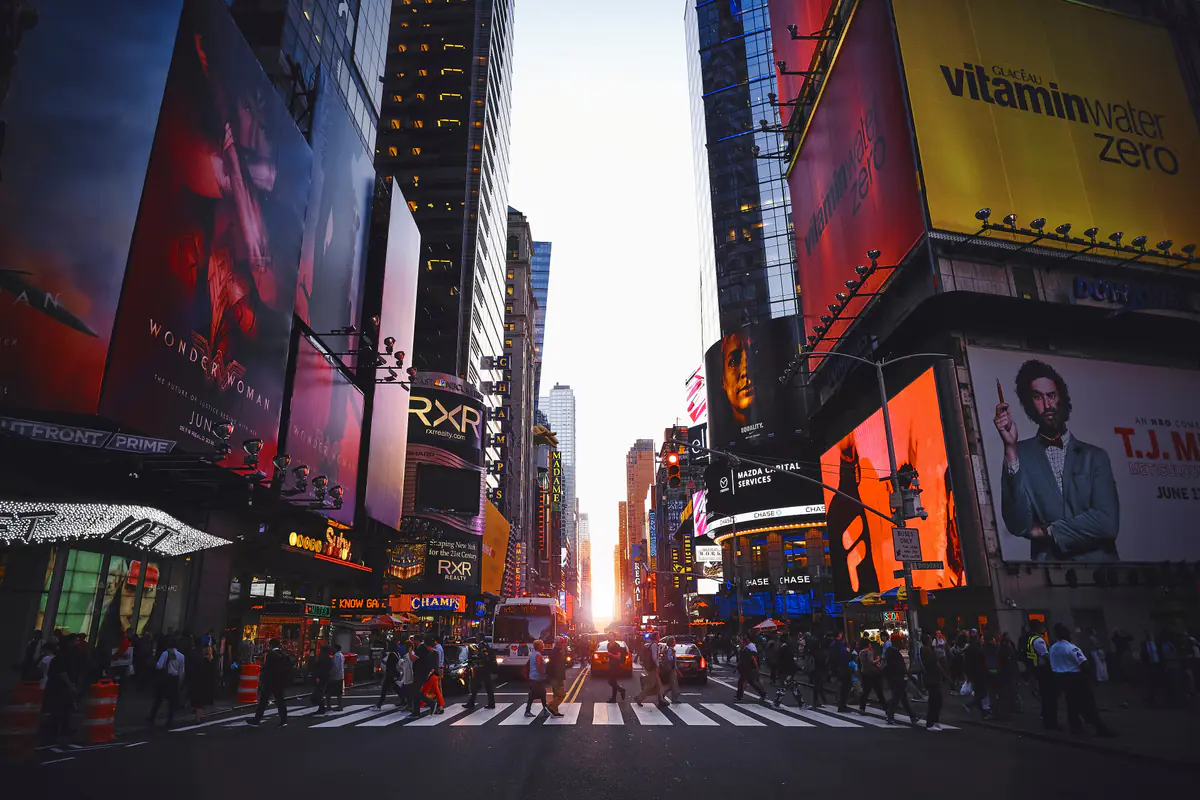 Times Square at sunset, crowds crossing the street between towers covered in lit billboards